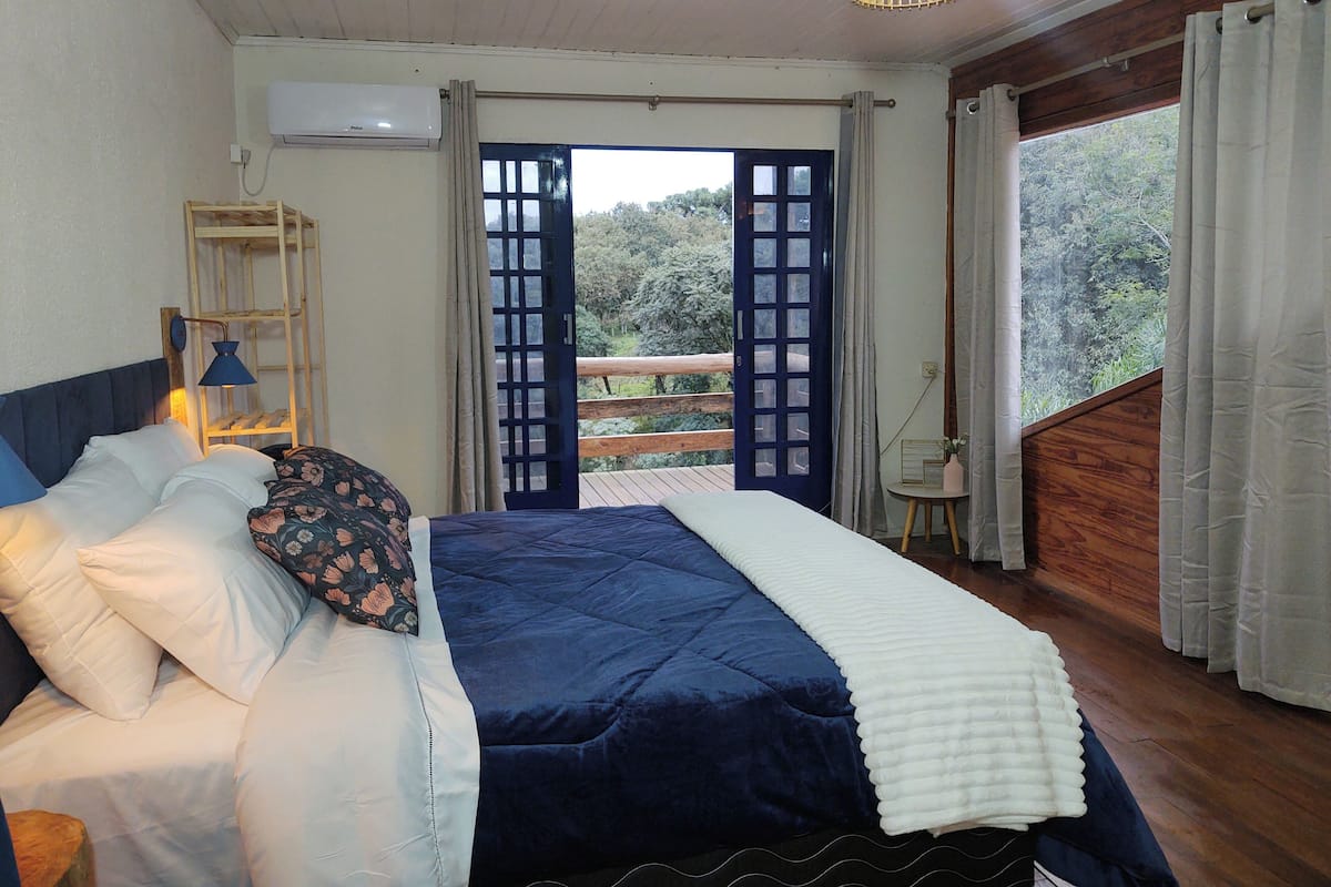 A bedroom with a bed, a bedside table, a lamp, a window with curtains, and a view of greenery outside.