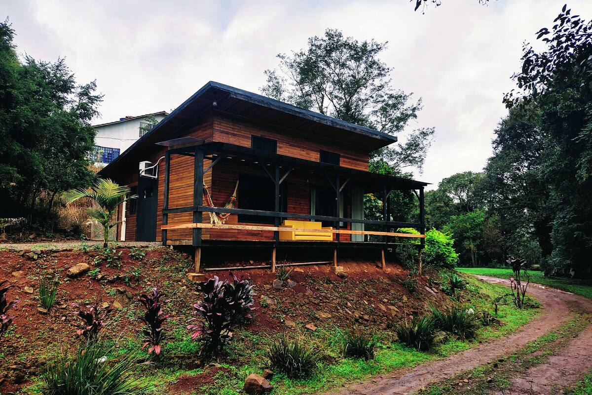 A wooden cabin with a porch, surrounded by lush greenery and a dirt path.