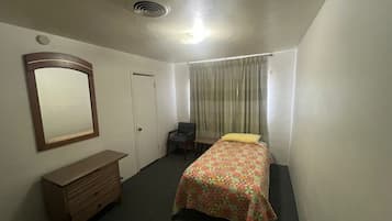 A small, single-bed room with a wooden dresser, a mirror, a door, and a window with curtains.