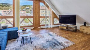 A modern living room with a large TV, wooden flooring, a blue sofa, a small table with fruit, and large windows offering a view of buildings and trees.