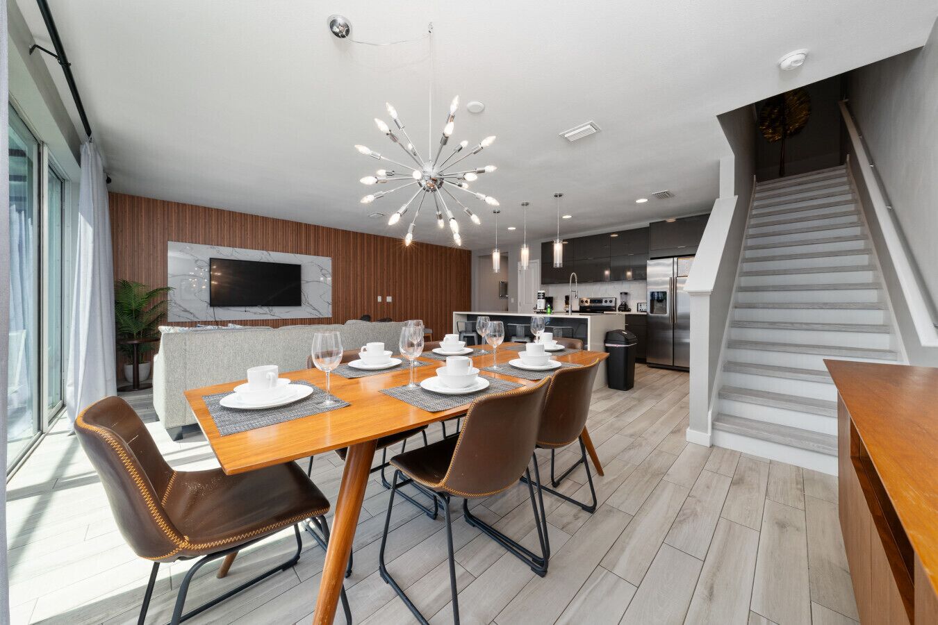 A modern dining area with a wooden table set for four, a large chandelier, and a kitchen in the background.