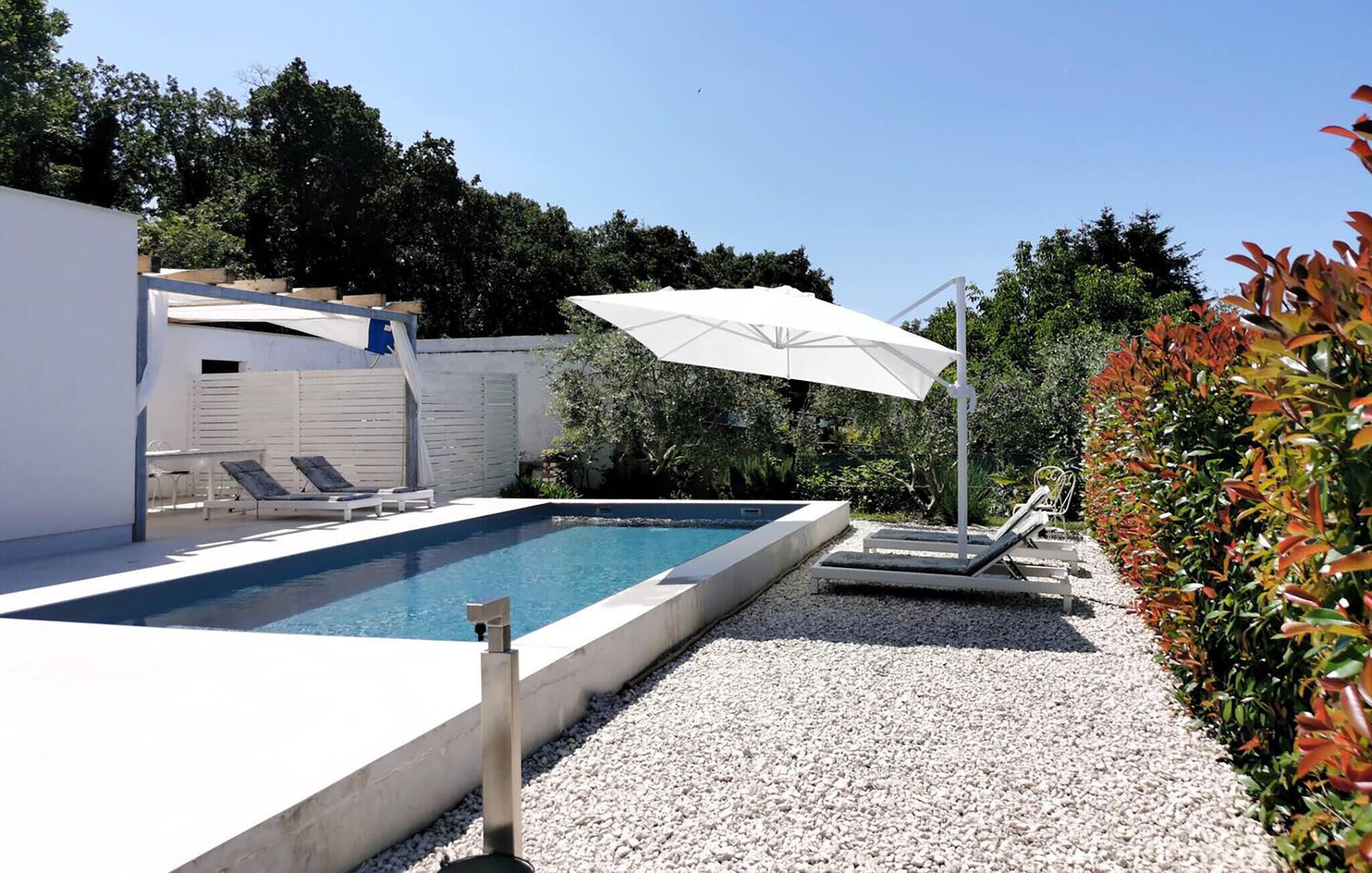 A modern outdoor swimming pool with a white deck, lounge chairs, and a pergola with a white umbrella.