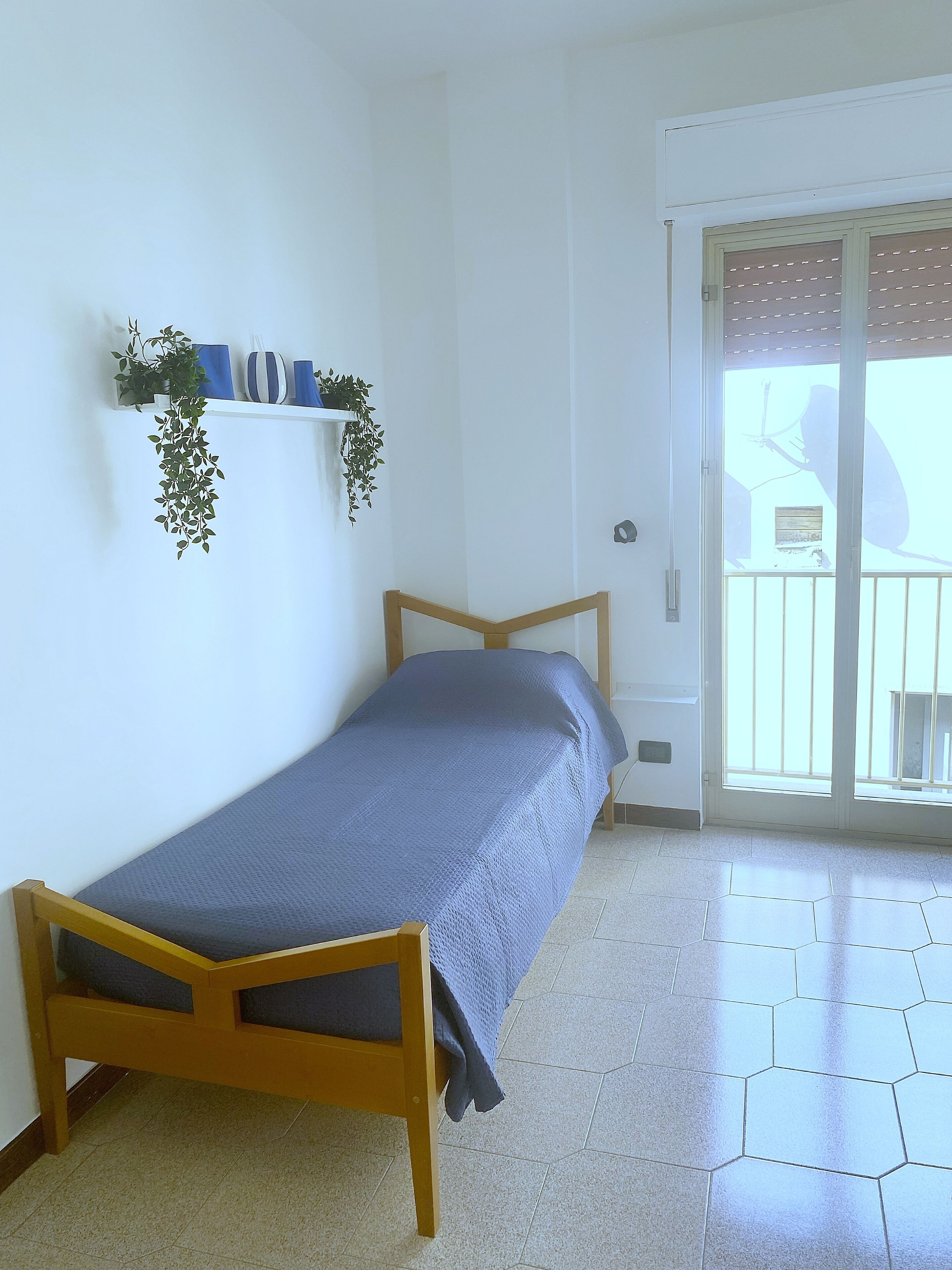 A small, single-bed room with a wooden frame, a blue bedspread, and a white tiled floor. There is a sliding glass door leading to an outdoor area with a balcony.