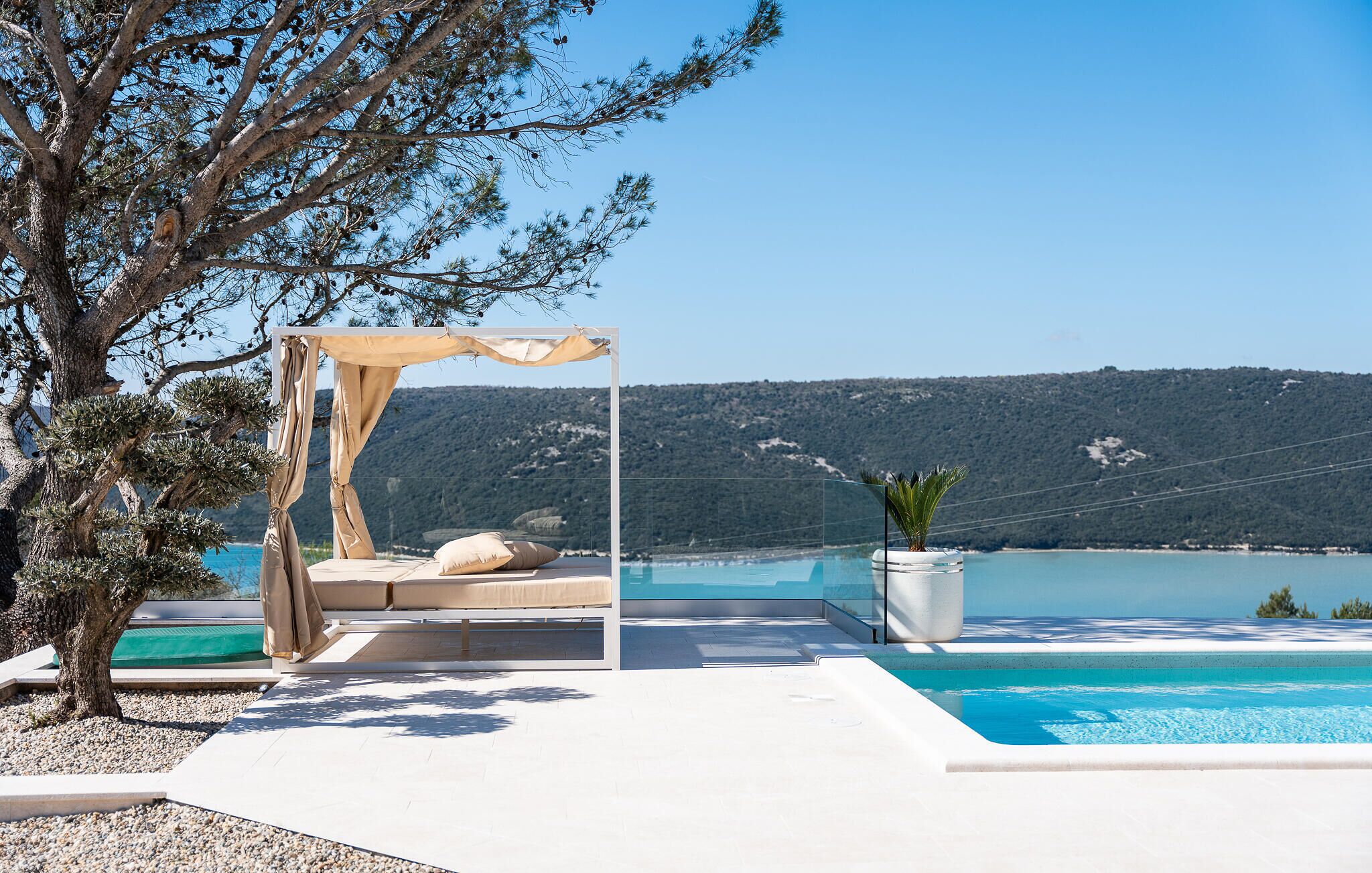 Un espace piscine privé doté d’un coin salon ombragé, offrant une vue sur un lac aux eaux d’un bleu limpide et sur les collines environnantes.