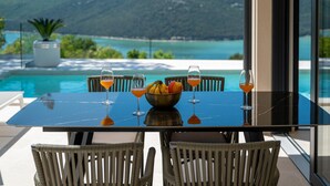 A table with four chairs, a bowl of fruit, and three glasses of orange juice, overlooking a pool and a scenic view of a mountain and sea. - Luxurious home in Trget (Trget)