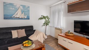 A modern living room with a black sofa, a wooden coffee table with a fruit bowl, a large painting of sailboats, a potted plant, a TV, and a wooden cabinet. - Lovely home in Selce with WiFi (Selce)