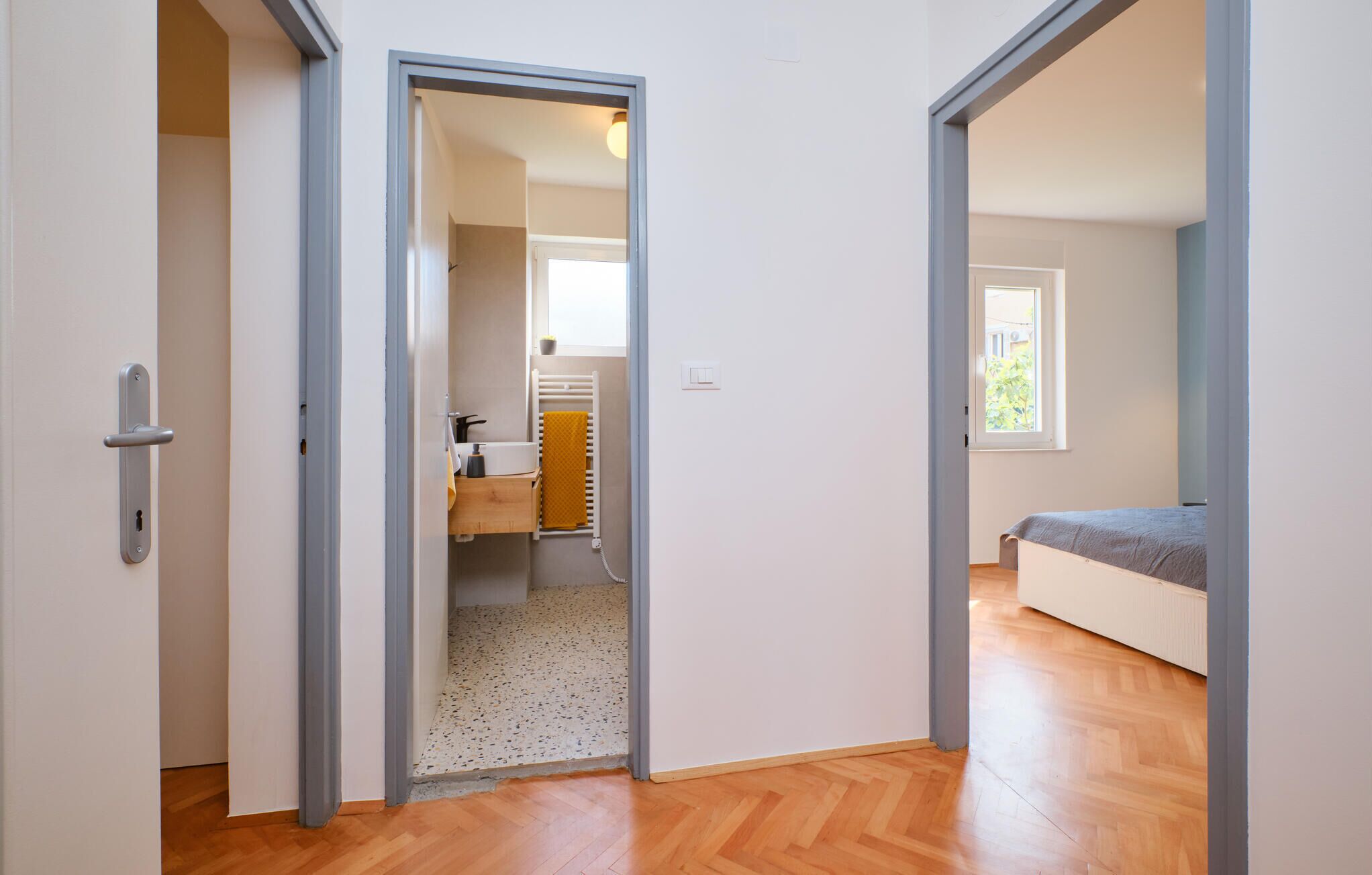 A hallway leading to a bathroom with a toilet, sink, and towel rack. There is a bed with a gray blanket in the adjacent room.
