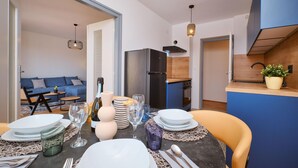 Ein Essbereich mit Tisch für vier Personen, eine Küche im Hintergrund und ein Wohnzimmer mit blauem Sofa.