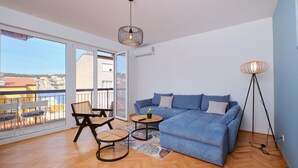 Ein modernes Wohnzimmer mit einem blauen Sofa, zwei Sesseln, einem kleinen Tisch und einer Stehlampe. Ein großes Fenster bietet Blick auf Gebäude und einen Balkon.