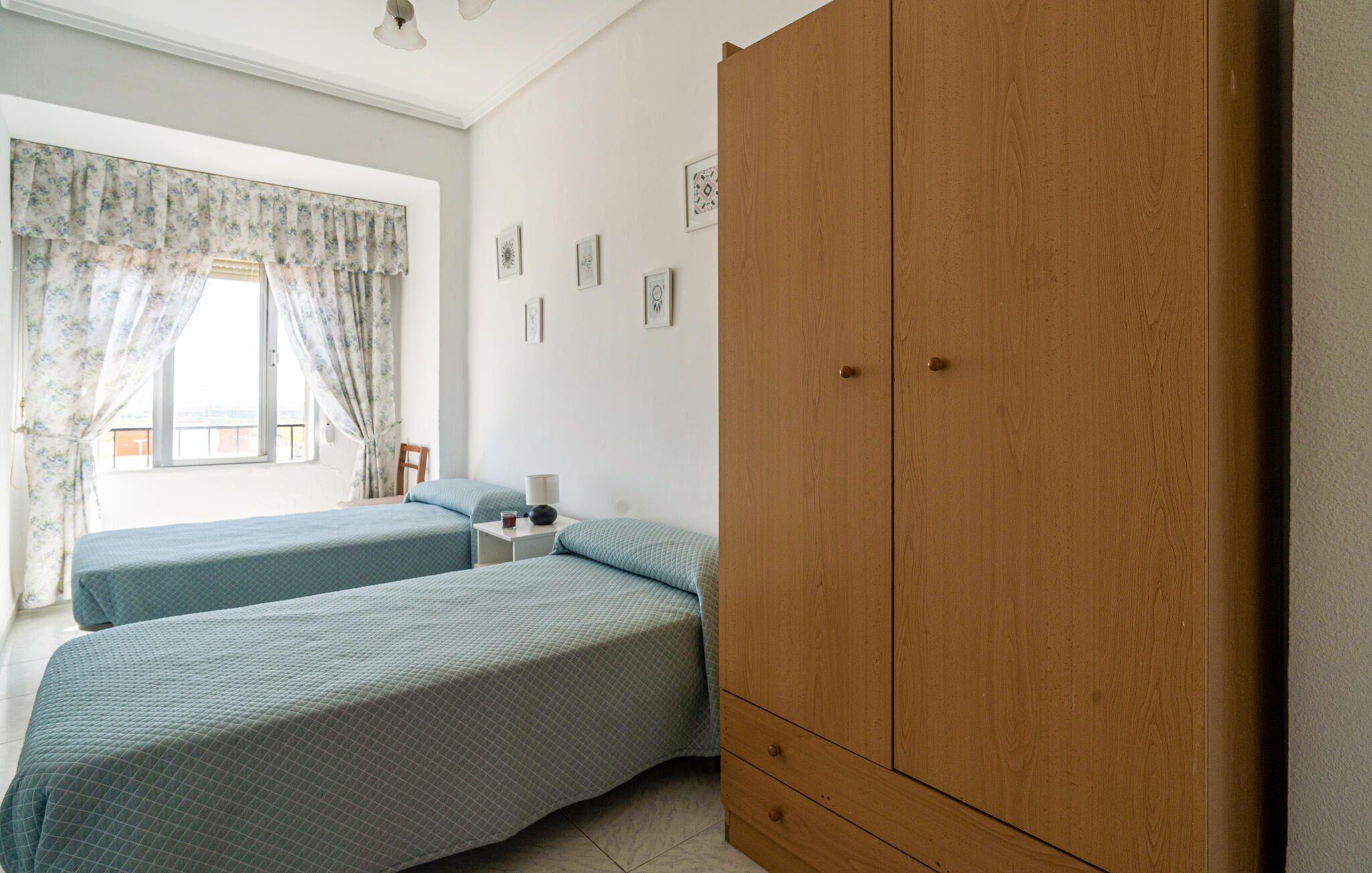 Een slaapkamer met twee eenpersoonsbedden, een houten garderobe, een nachtkastje en een raam met bloemen gordijnen.