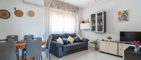 A modern living room with a grey sofa, dining table with chairs, a TV, and a cabinet with glass doors.