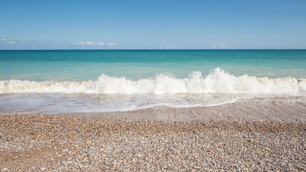 Una spiaggia ciottolosa con le onde che si infrangono sulla riva e un cielo azzurro limpido.