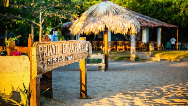 A wooden sign indicating a private beach club with a thatched roof structure in the background.