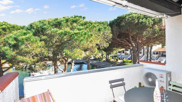 A balcony with a view of trees and a body of water, a chair, a table, and an air conditioning unit. - Beach front home in Saint-Cyprien Plage (Saint-Cyprien Plage)
