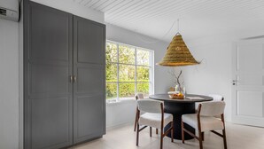 Een eetruimte met een ronde tafel, twee stoelen, een grote grijze garderobe, een raam met uitzicht op groen, en een hangende lamp.