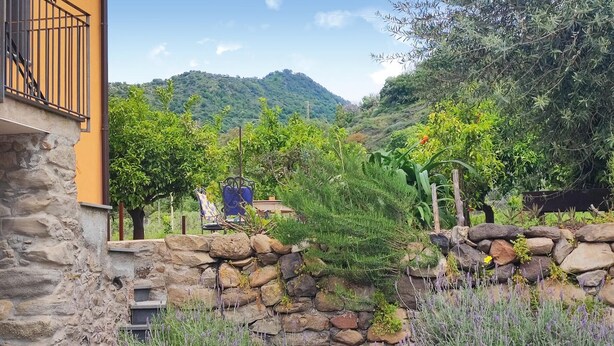 Un mur de pierre avec un jardin devant, un bâtiment jaune doté d’un balcon, et une montagne en arrière-plan.