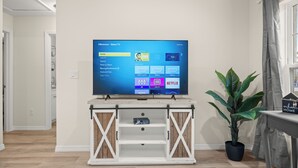 Olohuoneessa on seinälle asennettu televisio, jossa näkyy suoratoistopalvelun käyttöliittymä, valkoinen mediakeskus, ruukussa oleva huonekasvi ja ikkuna, jossa on verhot.