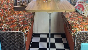 Et hyggeligt rum med et træbord, to mønstrede sofaer, en vintage radio og et fjernsyn på en tapetseret væg.