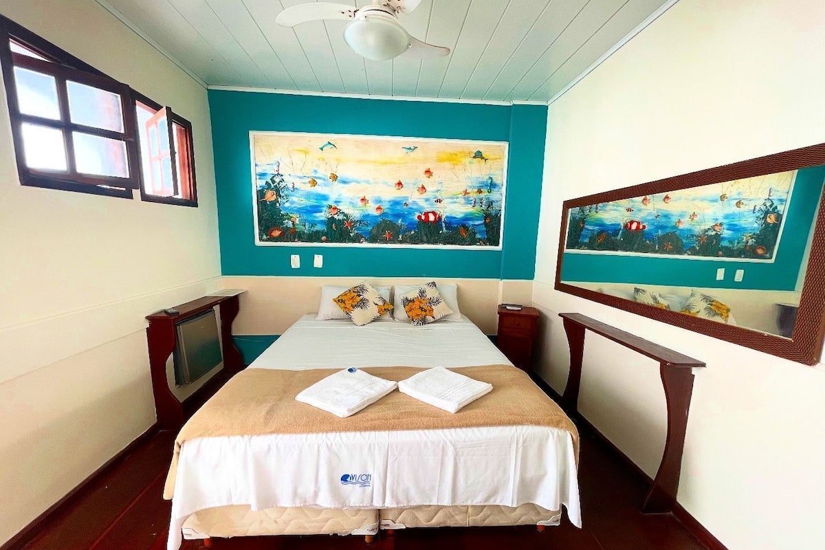 A hotel room with a bed, two bedside tables, a ceiling fan, and two paintings on the walls.