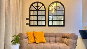A beige sofa with two bright orange cushions, a glass coffee table with a magazine, and a potted plant. Two arched windows with frosted glass panes are on the wall, and a hanging light fixture is visible.