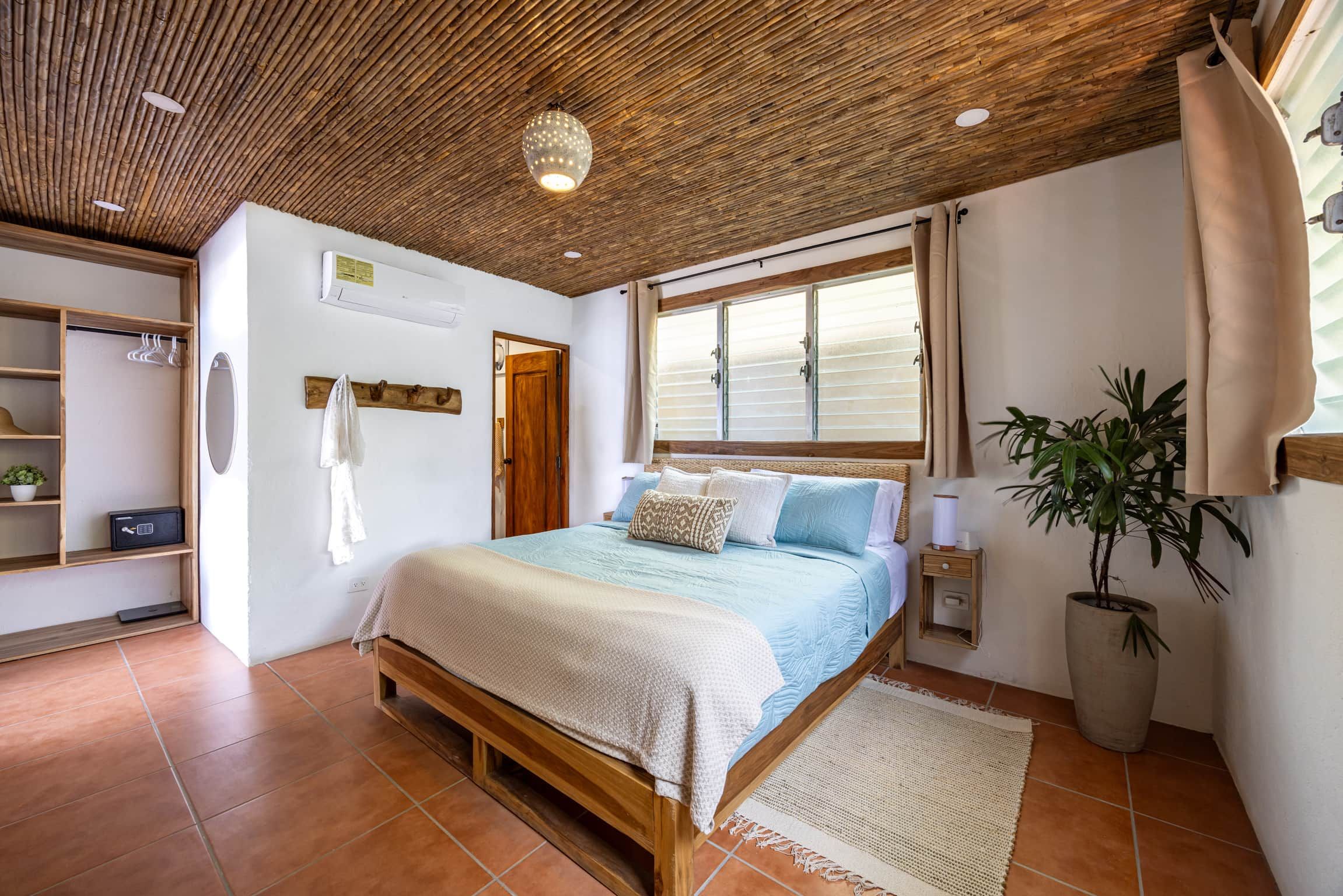 Une chambre avec un plafond en bois, un lit recouvert d’une couette bleue et blanche, une plante en pot et une fenêtre avec des stores.