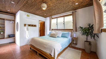A bedroom with a wooden ceiling, a bed with a blue and white duvet, a potted plant, and a window with blinds.