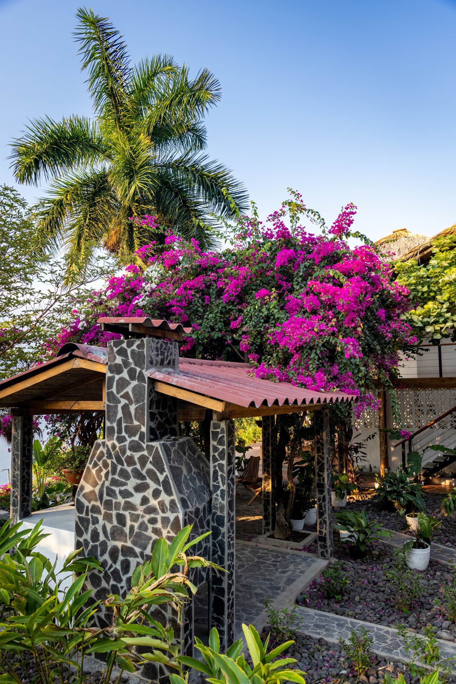 Estructura de piedra con tejado de baldosas, rodeada de exuberante follaje y vibrantes flores rosadas.