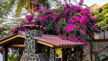 Estructura de piedra con tejado de baldosas, rodeada de exuberante follaje y vibrantes flores rosadas.