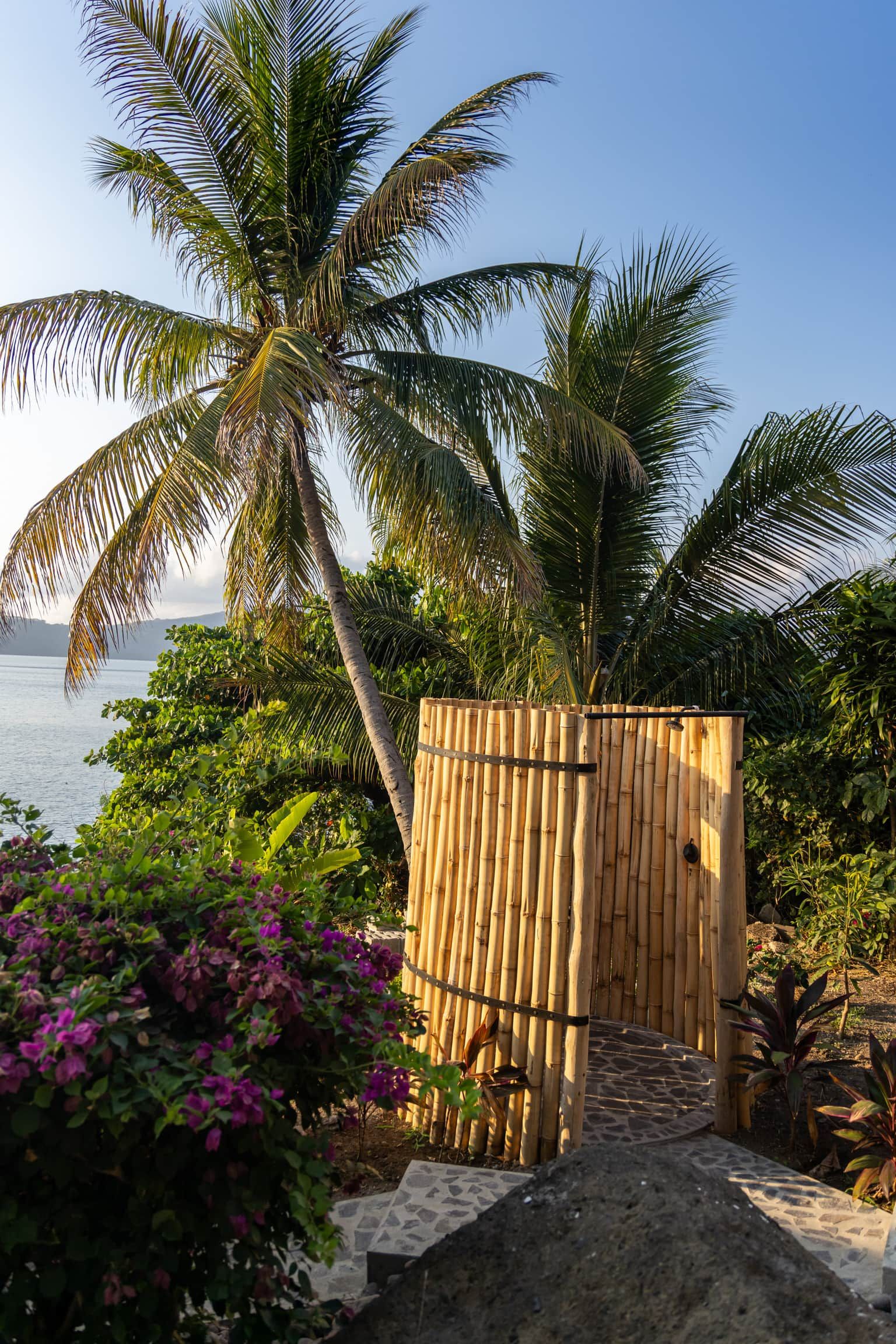 A bamboo gate leading to a beach area with palm trees and purple flowers.