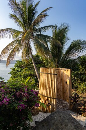 Una puerta de bambú que da a una zona de playa con palmeras y flores púrpuras.