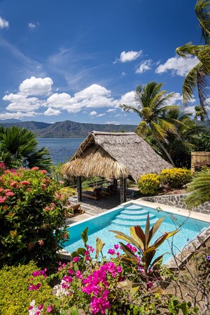 Un área de piscina con una estructura de techo de paja, rodeada de plantas y flores tropicales.