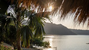 A beachside lounge with wooden loungers and a palm tree, overlooking a calm sea and a mountainous backdrop.