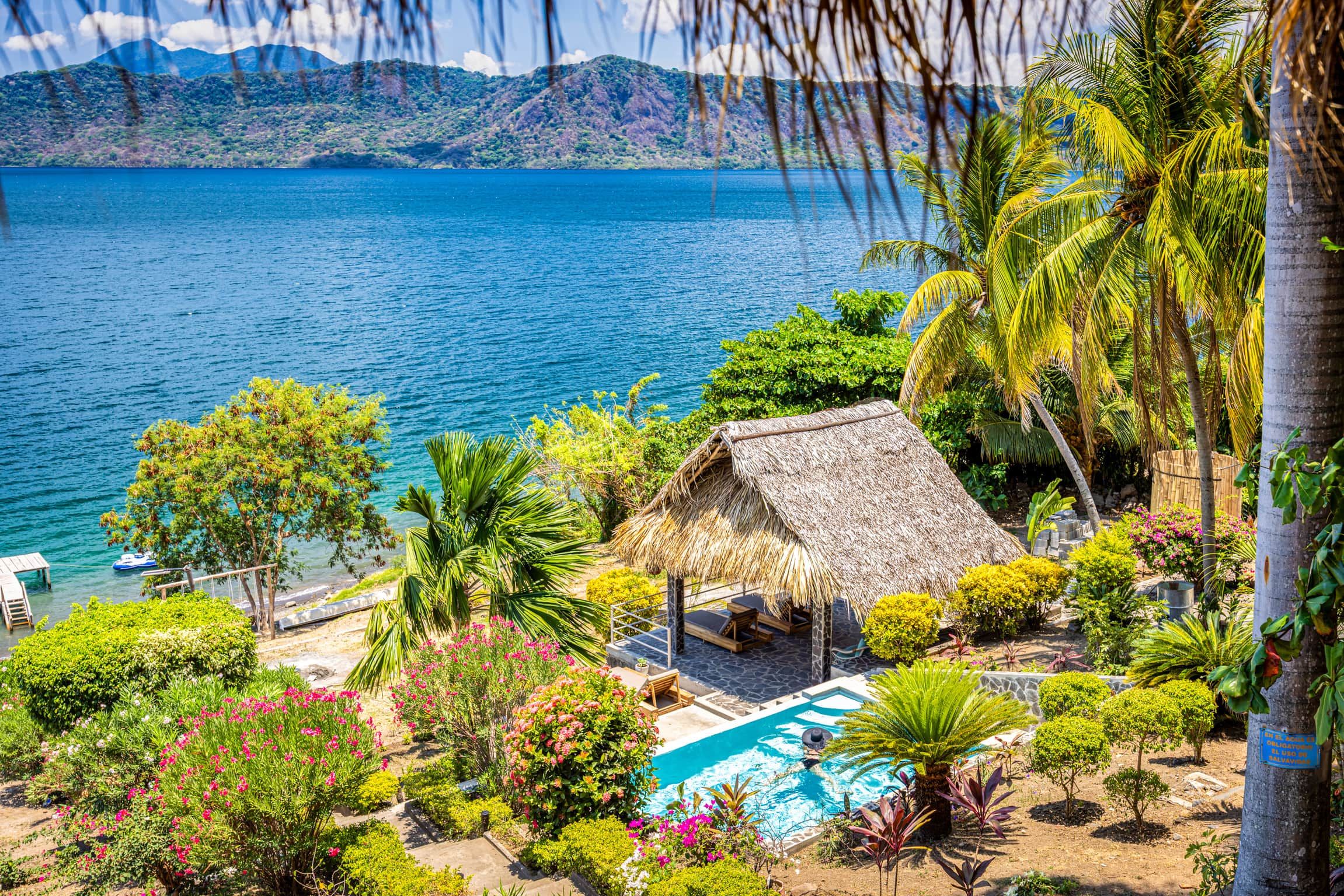 Un resort tropical con piscina, construcción de techo de paja y exuberante follaje.