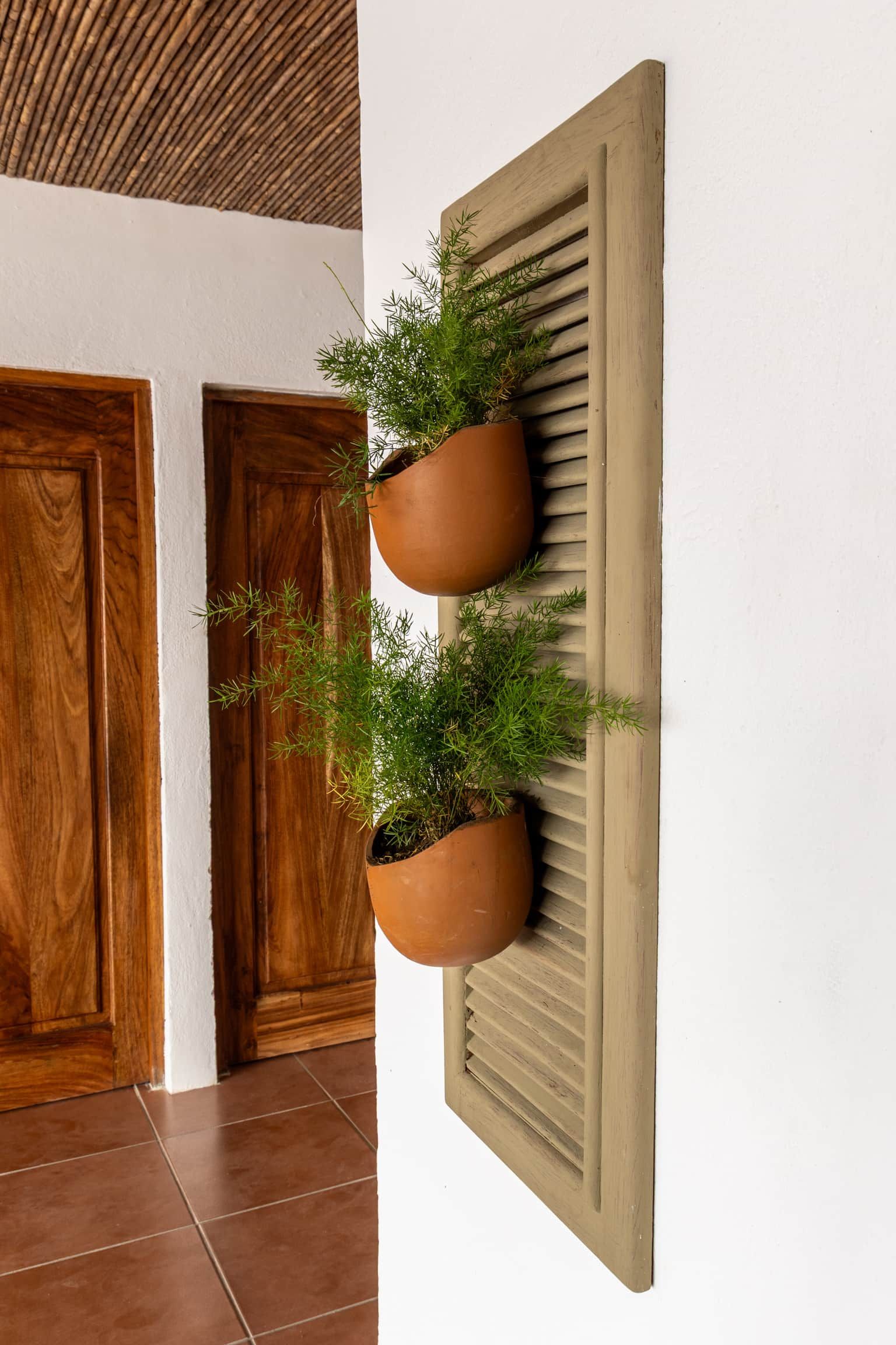 A wall-mounted planter with two terracotta pots containing greenery, positioned between two wooden doors in a room with a tiled floor.
