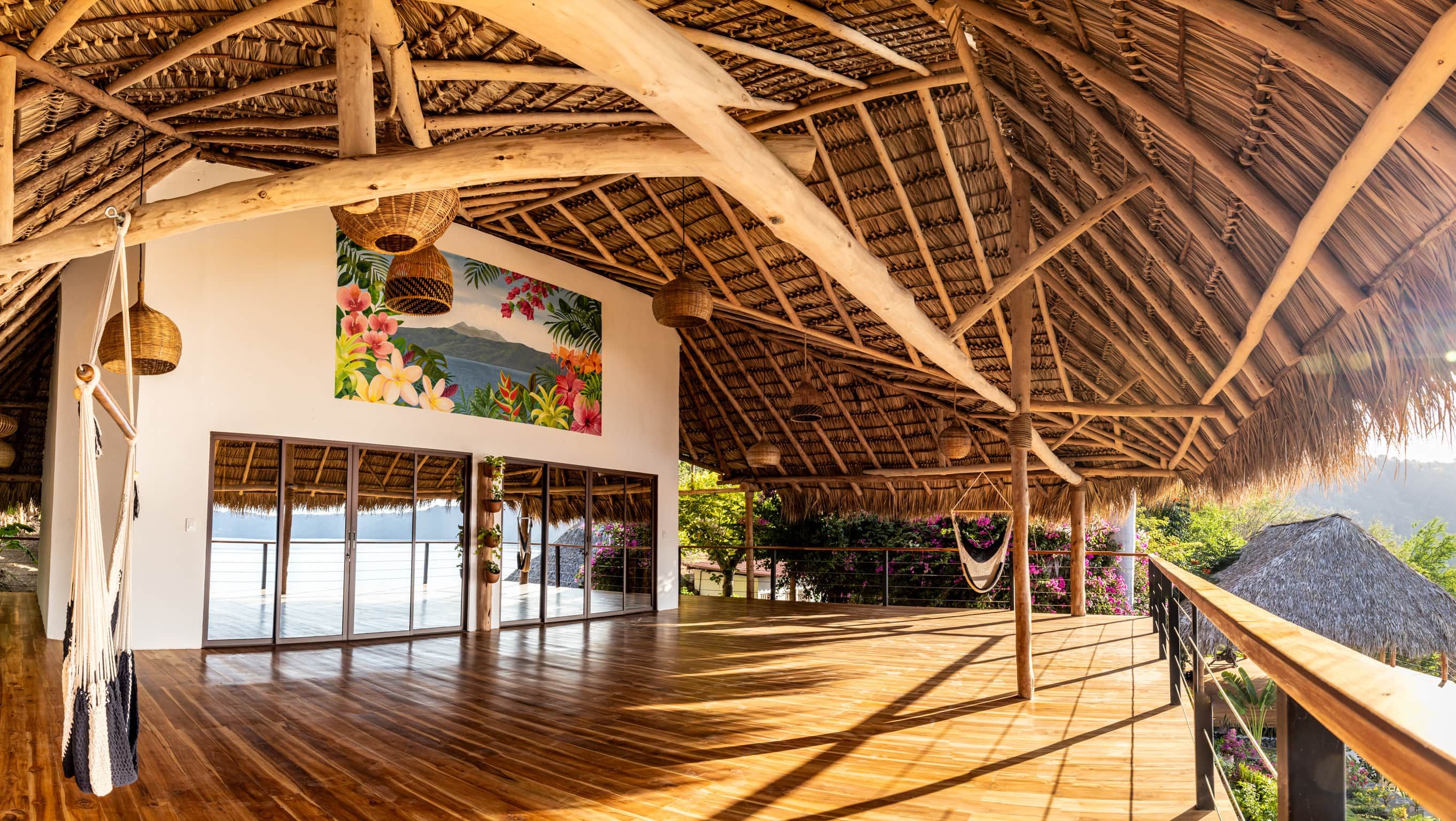 A spacious room with a wooden floor, large windows, and a thatched roof.