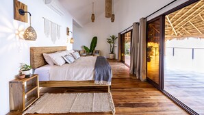A spacious bedroom with a large bed, wooden floor, and a view of a thatched-roof structure through a sliding glass door.