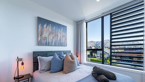 Un dormitorio moderno con una cama grande, un cuadro en la pared y vistas a un pueblo a través de amplios ventanales.