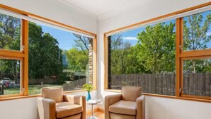 Camera d'angolo con due poltrone beige, un tavolino bianco e ampie finestre che danno su un giardino con alberi e una recinzione in legno.