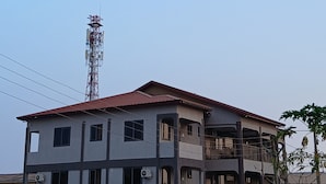 Un edificio de varios pisos con techo rojo, varias ventanas y una torre de telecomunicaciones al fondo.