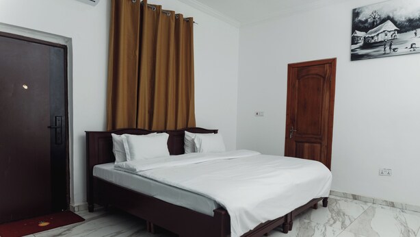 A bedroom with a wooden bed, white bedding, a brown curtain, a wooden door, and a painting on the wall. - RAGANT HOTEL (Cape Coast)