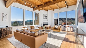 A spacious living room with a large brown leather sofa, wooden beams on the ceiling, and large windows offering a view of the mountains. - The Belvedere // Hot Tub, Sauna & Red Rock Views (Sedona)