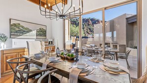 A dining area with a large wooden table set for six, surrounded by chairs. There is a chandelier above the table, and a painting of a landscape on the wall. - The Belvedere // Hot Tub, Sauna & Red Rock Views (Sedona)