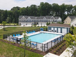 A rectangular swimming pool with blue lounge chairs and a fenced deck area, surrounded by a residential neighborhood with houses.