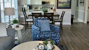A spacious living room with a round coffee table, blue patterned armchairs, a dining area with a chandelier, and a kitchen in the background. - Callaway Corner | Luxe Pet-Friendly 2BR Condo (Myrtle Beach)