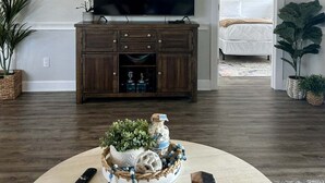A living room with a TV, wooden cabinet, potted plants, and a view of the beach through a door. - Callaway Corner | Luxe Pet-Friendly 2BR Condo (Myrtle Beach)