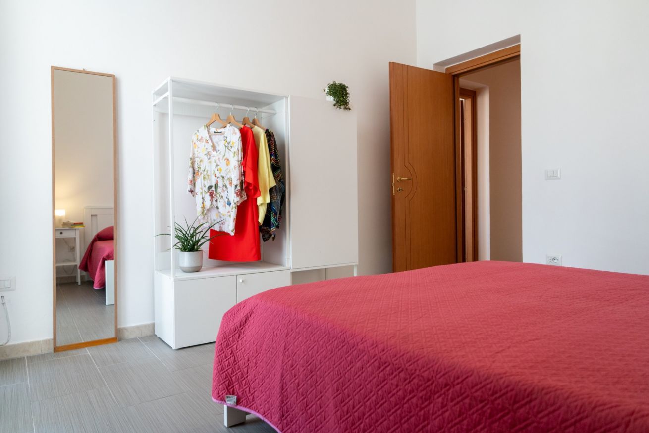 Ein Schlafzimmer mit roter Bettdecke, einem weißen Kleiderschrank mit darinhängender Kleidung und einer Holztür.