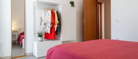 A bedroom with a red bedspread, a white wardrobe with hanging clothes, and a wooden door.