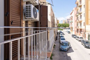 A residential street with parked cars, a balcony with a metal railing, and buildings with closed shutters.