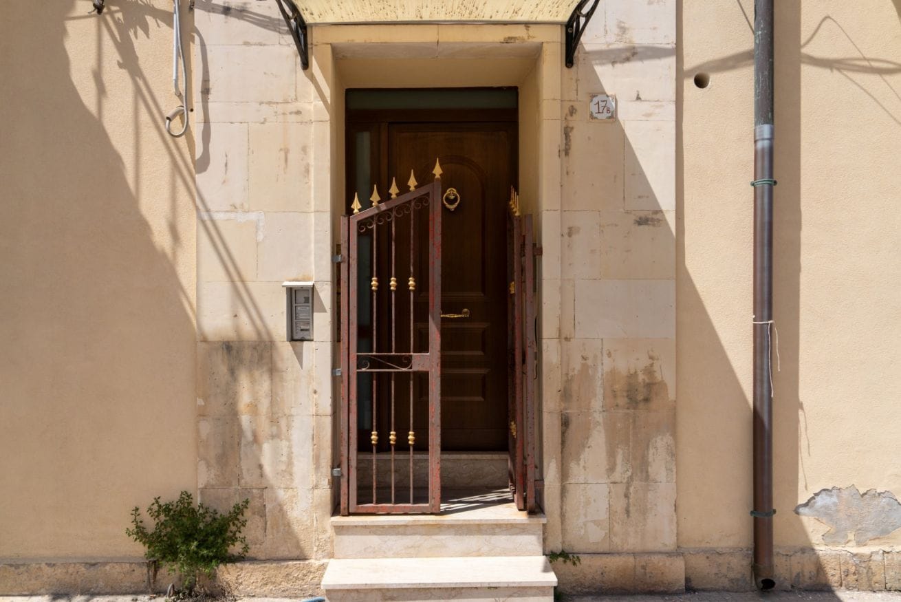 A beige building with a wooden door, a small step leading up to the entrance, and a metal pipe on the right.