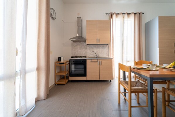 A compact kitchen with a gas stove, oven, and a small wooden shelf. There is a dining table with chairs and a window with curtains.